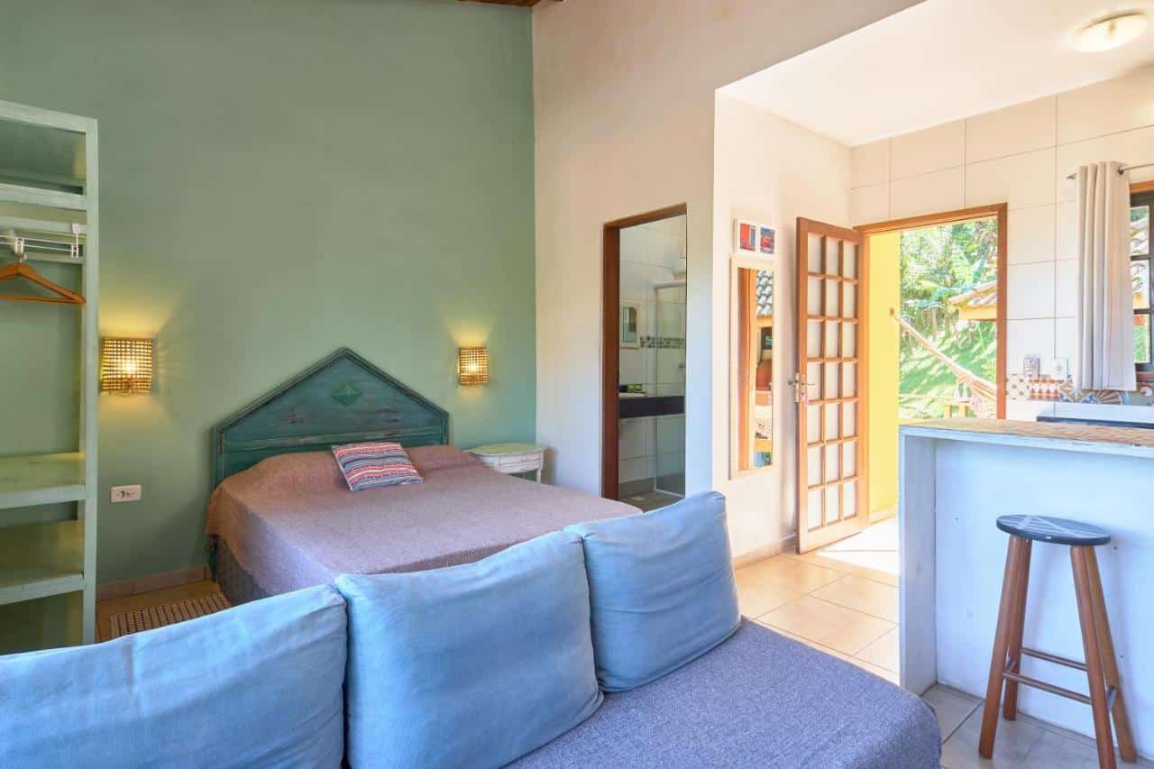 Um quarto com uma parede azul-petróleo, cama, prateleiras e uma porta aberta que leva a uma sacada. Um sofá e um banco de bar estão no primeiro plano.