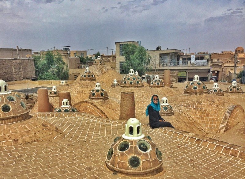 Uma pessoa sentada em um telhado de tijolos tradicional com cúpula e aberturas circulares de vidro, cercada por um céu nublado e edifícios no Sultan Amir Ahmad Bathhouse.
