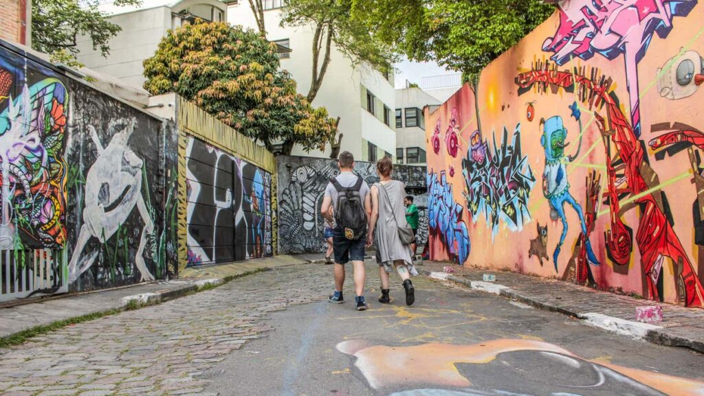 Um grupo de pessoas caminha na Vila Madalena por uma rua de paralelepípedos ladeada por muros coloridos cobertos de grafite e árvores no alto.