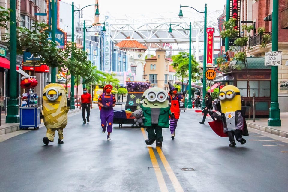 Cinco pessoas fantasiadas de Minions, algumas usando fantasias de Halloween, caminham pela rua de um parque de diversões com prédios coloridos.