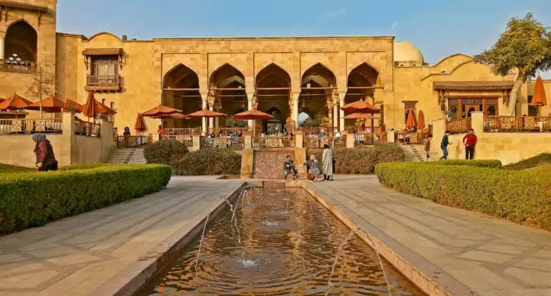 Edifício de pedra com entradas em arco, assentos ao ar livre e uma fonte central cercada por sebes e pessoas na Al-Azhar Park, que pode ser visitada com eSIM Cairo.