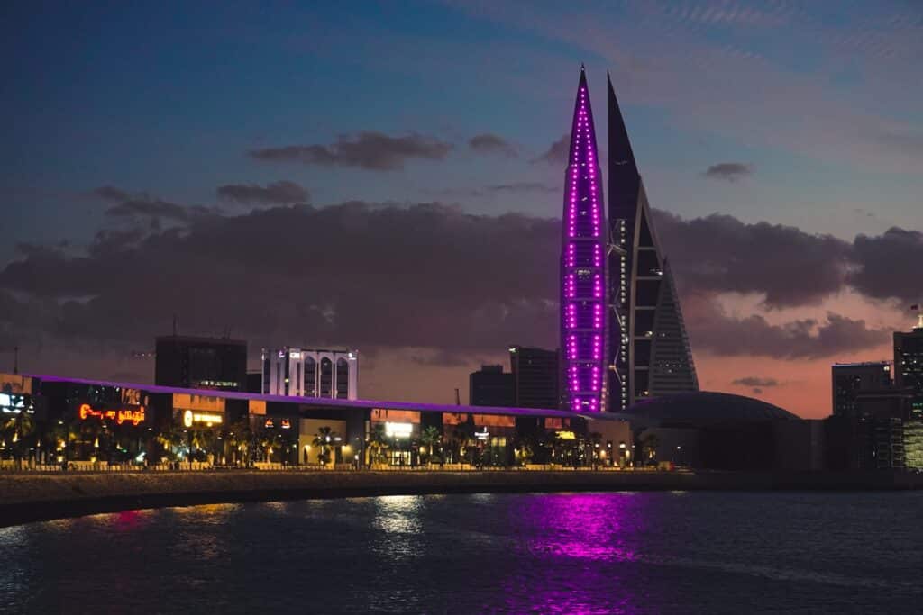 Bahrain World Trade Center, Manama em um horizonte da cidade ao anoitecer com dois prédios altos iluminados por luzes roxas refletidas na água, com céu nublado acima, que pode ser visitada com o eSIM Bahrein.