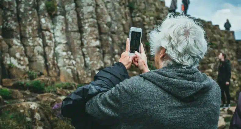 Pessoa com cabelos grisalhos tira uma foto com um smartphone perto de grandes formações rochosas; outras pessoas estão ao fundo nas Falésias de Moher, que pode ser visitada com o eSIM Irlanda.