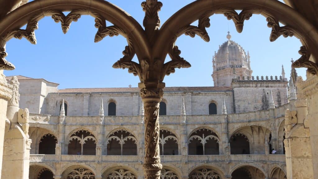 Arcos de pedra emolduram a vista do Mosteiro dos Jerônimos, um claustro histórico ornamentado com entalhes detalhados e uma alta torre abobadada sob um céu claro, que pode ser observado com o eSIM Lisboa.