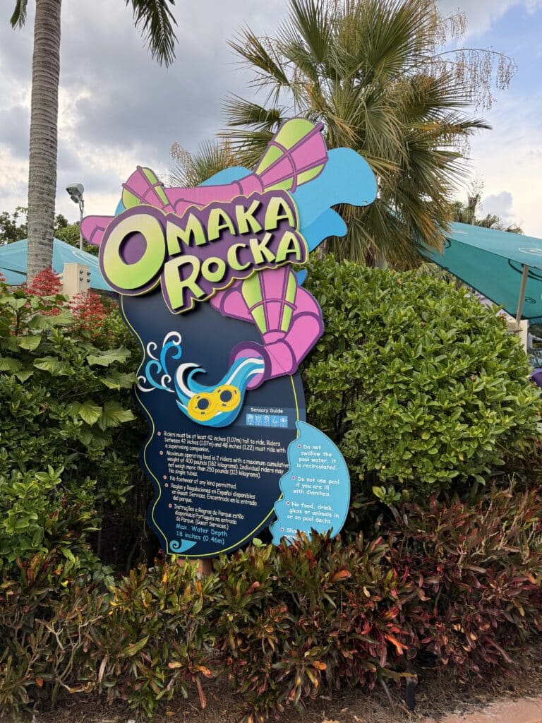 Placa colorida do toboágua "Omaka Rocka" cercada por arbustos verdes, palmeiras e um céu azul ao fundo.