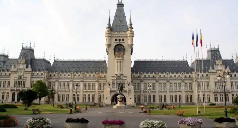 O Palácio da Cultura em Iași, um edifício grande e ornamentado com uma torre de relógio central, canteiros de flores, bancos e três mastros de bandeira na frente, que pode ser visitado com o eSIM Romênia.
