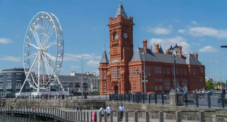 Um grande edifício de tijolos vermelhos e uma roda-gigante erguem-se à beira-mar sob um céu azul claro. Pessoas caminham ao longo do cais em Pierhead Building, Cardiff, que pode ser visitada com o eSIM País de Gales.