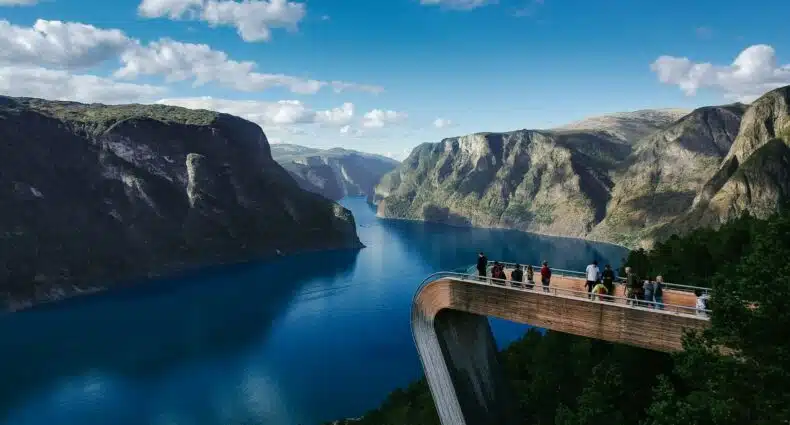 Pessoas ficam em uma plataforma de observação moderna com vista para um fiorde azul cercado por montanhas íngremes sob um céu parcialmente nublado em Stegastein, Aurland que pode ser visitada com o eSIM Noruega.