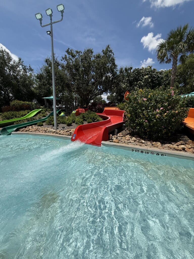 Um toboágua vermelho no Turi's Kid Cove termina em uma piscina clara, cercada por vegetação e outros toboáguas sob um céu ensolarado no Aquatica Orlando.