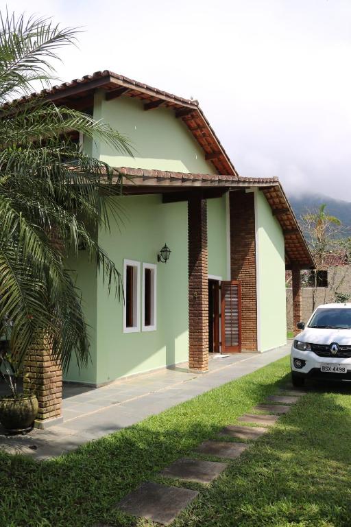 Uma casa verde-clara com detalhes em tijolos, um telhado de telhas e um carro branco estacionado em um caminho de pedra ao lado de um gramado, trata-se da propriedade Aconchegante Casa de Temporada.