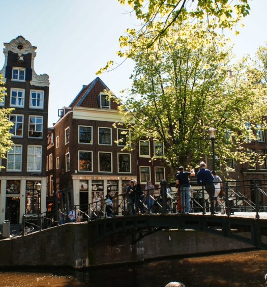 Pessoas em uma ponte sobre um canal, com edifícios históricos e árvores ao fundo em um dia ensolarado em Amsterdam que pode ser visitada com o eSIM Holanda.