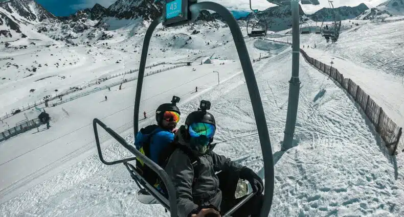Duas pessoas com equipamento de esqui andam de teleférico em Pas de la Casa, Andorra, por uma encosta nevada de montanha, tirando uma selfie com um bastão de selfie. A foto é capa do post de eSIM Andorra.