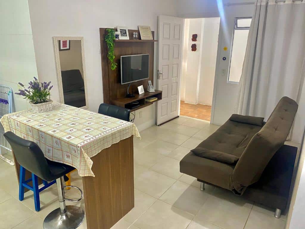 Pequena sala de estar com um sofá marrom, TV em um suporte, mesa de jantar com uma toalha de mesa, duas banquetas e um espelho de parede, no Apartamento Floripa 16.