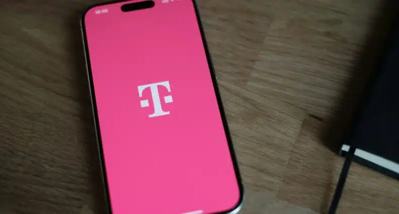 Um smartphone exibe o logotipo da T-Mobile em uma tela rosa, sobre uma superfície de madeira ao lado de um notebook preto. A imagem ilustra o post sobre eSIM T-Mobile.