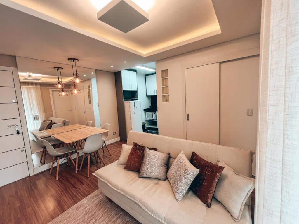 Sala de estar moderna com um sofá bege, almofadas estampadas, mesa de jantar e vista para uma área de cozinha compacta, no Apto conforto extremo.