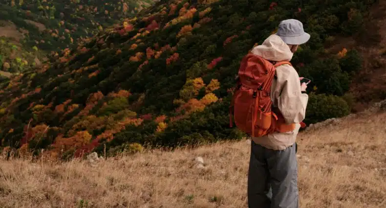 Uma pessoa com uma mochila laranja está em uma colina gramada na Armênia, olhando para um telefone, com uma encosta florestal colorida ao fundo. A foto é a capa do post de eSIM Armênia.