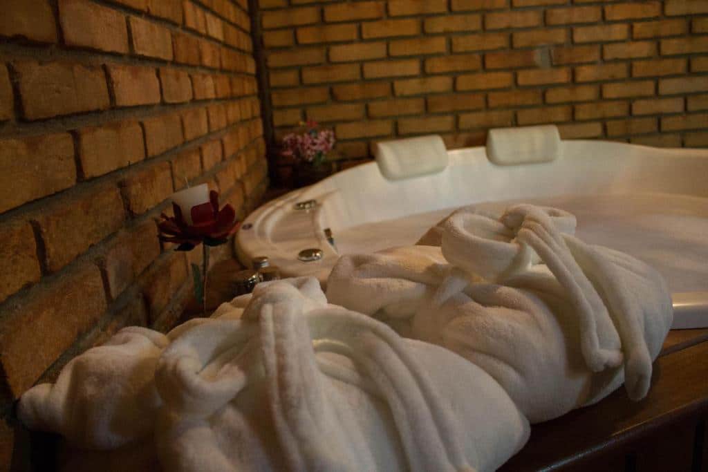 Dois roupões de banho brancos estão dispostos em frente a uma banheira de hidromassagem com dois apoios de cabeça, encostados em uma parede de tijolos, em um quarto na Pousada da Carmem.