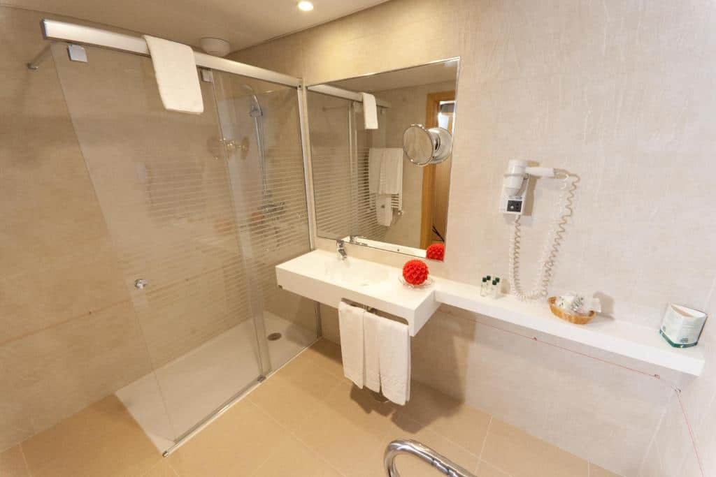 Banheiro moderno com chuveiro de vidro, pia branca, toalhas, secador de cabelo na parede e produtos de higiene pessoal no balcão, na propriedade do Hotel Cinquentenário & Conference Center.