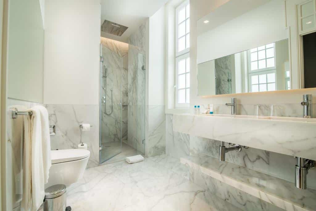 Banheiro moderno com piso e paredes de mármore, pia dupla, espelho grande, vaso sanitário, toalheiro e chuveiro de vidro, em um dos quartos do Hotel da Baixa.