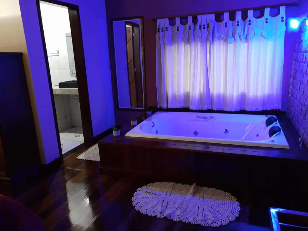 Grande banheira branca com jatos em uma estrutura de madeira, perto de uma janela com cortinas; a porta leva a um banheiro, no Sítio Recanto do Araçá em Visconde de Mauá.