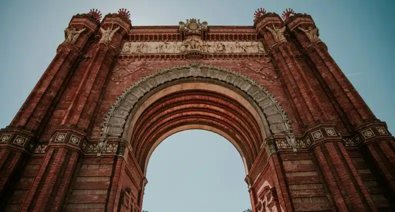 Vista de ângulo baixo de um grande arco de tijolos vermelhos com entalhes e colunas ornamentadas contra um céu azul claro. Ilustra o post sobre eSIM Barcelona.
