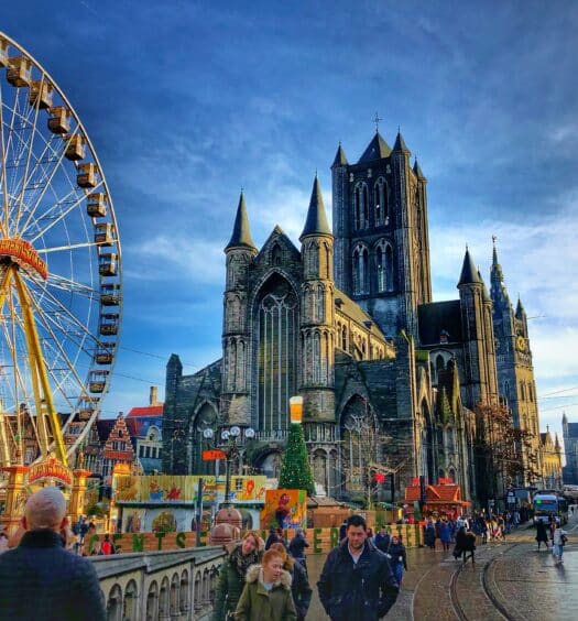 Uma grande igreja gótica fica ao lado de uma roda-gigante em uma movimentada praça da cidade de Gante, na Bélgica, com pessoas caminhando e edifícios próximos. A foto é capa do post de eSIM Bélgica.
