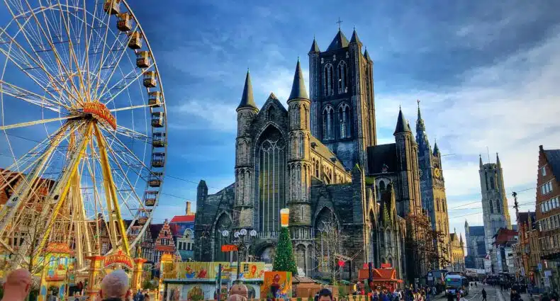Uma grande igreja gótica fica ao lado de uma roda-gigante em uma movimentada praça da cidade de Gante, na Bélgica, com pessoas caminhando e edifícios próximos. A foto é capa do post de eSIM Bélgica.