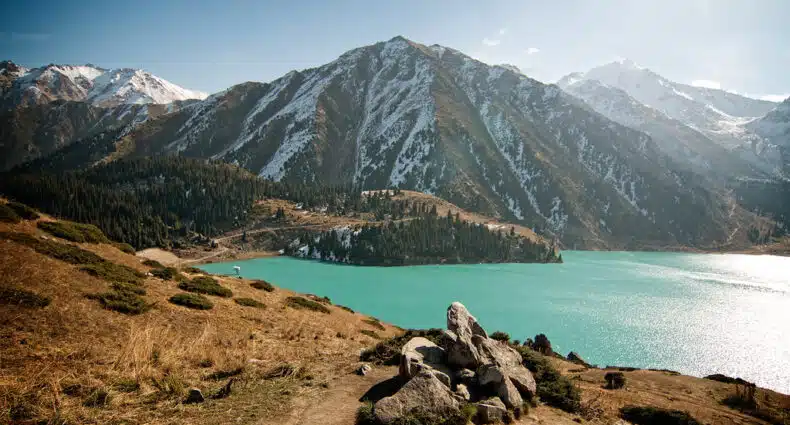 O Big Almaty Lake, um lago azul-turquesa fica na base de montanhas cobertas de neve sob um céu claro, com rochas em primeiro plano. Imagem para ilustrar post sobre eSIM Cazaquistão.