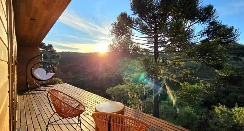 Deck de madeira com duas cadeiras laranja e uma pequena mesa, com vista para as árvores e colinas ao pôr do sol, nas Cabanas Alto Padrão com Jacuzzi, Lareira e cachoeira exclusiva, para representar pousadas em Urupema.