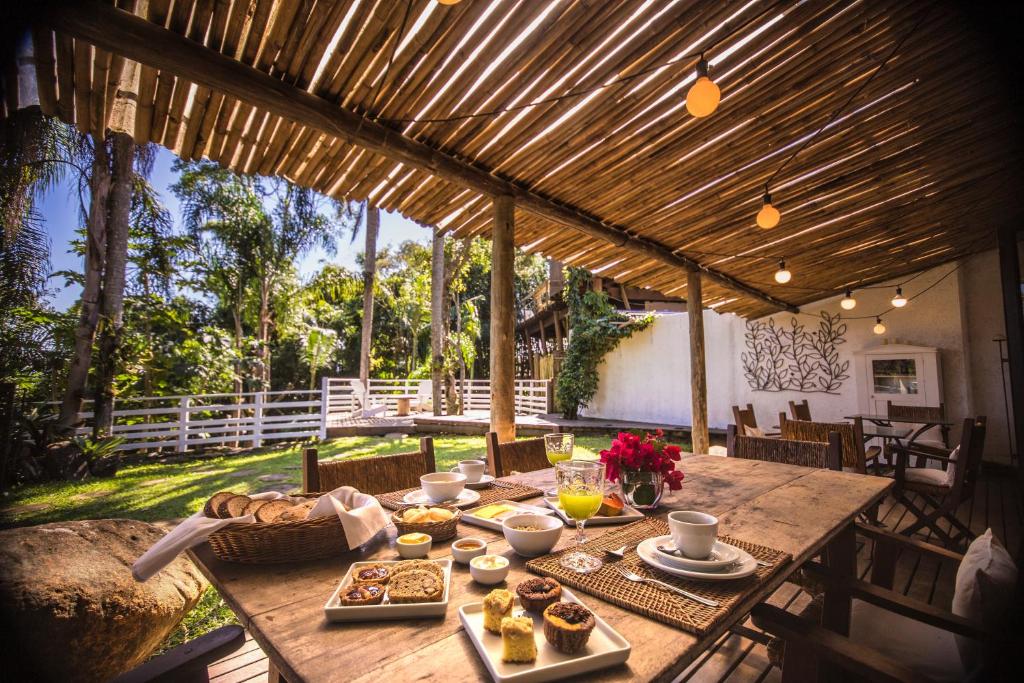 Uma mesa de jantar rústica ao ar livre é posta com alimentos para o café da manhã, bebidas e flores sob uma pérgola de madeira em um jardim, na propriedade da Hospedaria das Brisas.