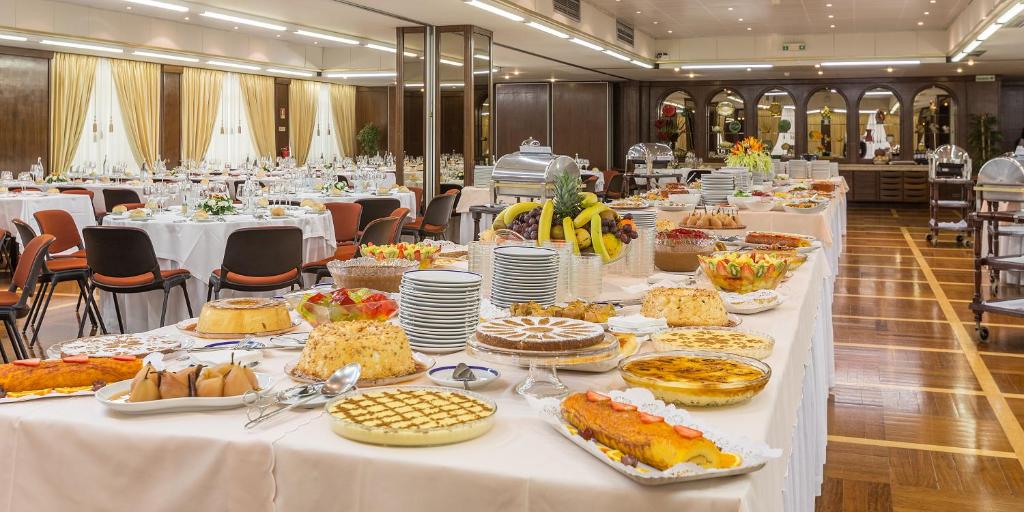 Uma longa mesa de bufê com sobremesas e frutas variadas é montada em um salão de banquetes formal com mesas de jantar redondas, servido no Hotel Fátima.