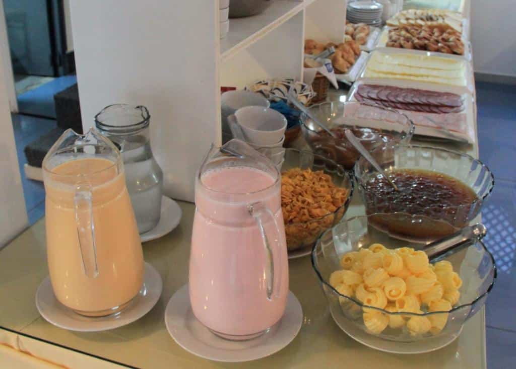 Um bufê de café da manhã com jarras de leite e suco, tigelas de cereal, geleia, manteiga, carnes e fatias de queijo, servido no Hotel Florinda.