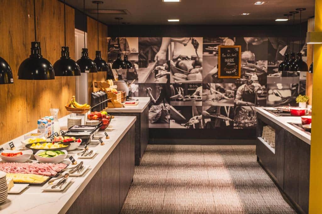 Buffet no ibis London Excel-Docklands com vários alimentos em dois balcões longos, incluindo frutas, queijo, carnes fatiadas, pães e cereais.