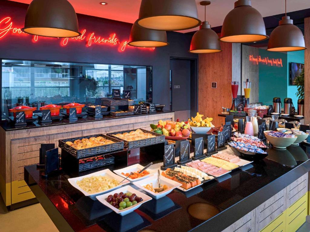 Um bufê de café-da-manhã com doces, frutas, queijos, frios, distribuidores de suco e iluminação moderna no alto, no ibis São Paulo Tatuape.