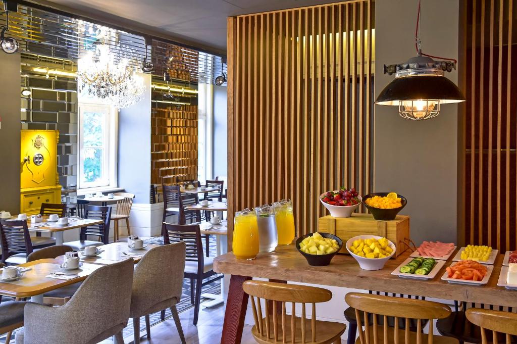 Um bufê de café da manhã com frutas e sucos fica sobre uma mesa de madeira em uma sala de jantar moderna e bem iluminada, com mesas e cadeiras vazias, no My Story Hotel Rossio.
