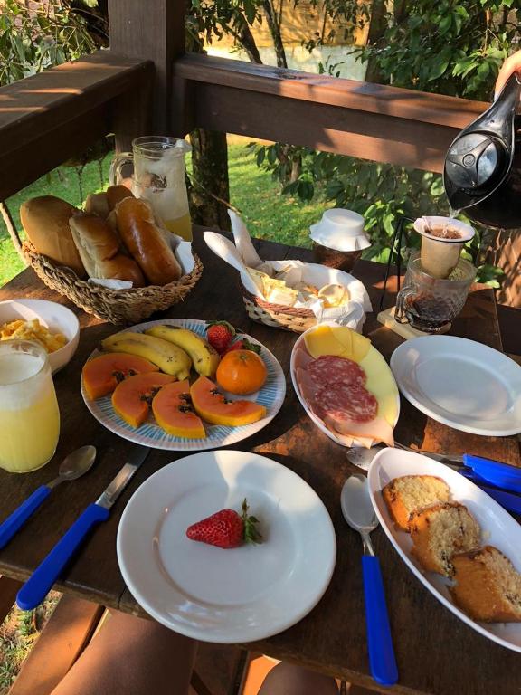 Uma mesa de madeira posta para o café da manhã com pães, frutas, suco, bolo, frios e café, ao ar livre, com vegetação, no Farol do Arvoredo Cabanas.