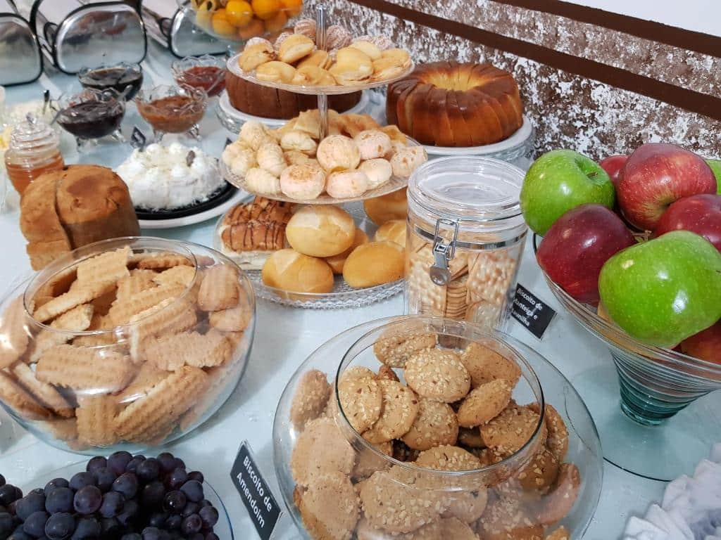 Uma mesa exibe uma variedade de produtos assados, pães frescos, biscoitos, maçãs, uvas e potes de alimentos em conserva, servido na Pousada Lugama.