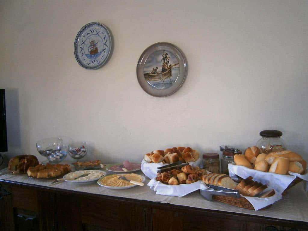 Uma mesa de bufê com pães variados, bolos, frios, queijos e potes, sob dois pratos decorativos na parede, servido na Pousada Mar e Marés.