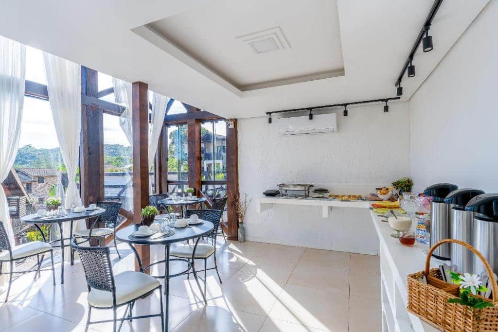 Área de jantar bem iluminada com mesas redondas, cadeiras, balcão de bufê e distribuidores de café perto de grandes janelas com cortinas transparentes, na Pousada Solar dos Lírios.