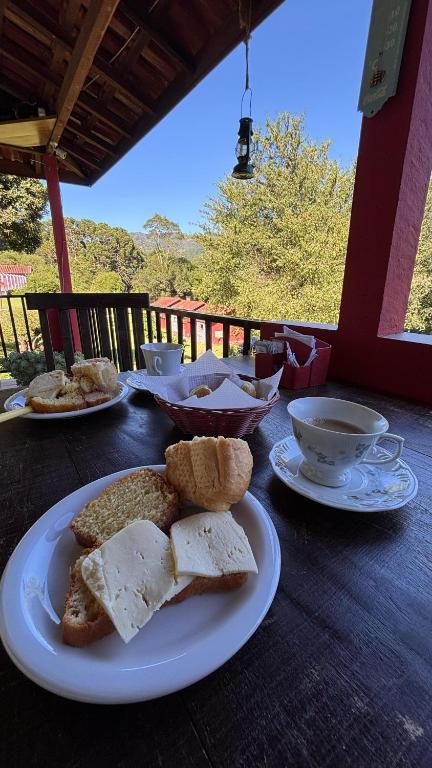 Uma mesa de madeira com café, pão, queijo e bolos em pratos, colocada em uma varanda com árvores e céu azul ao fundo, servido na propriedade do Residencial das Araucárias.