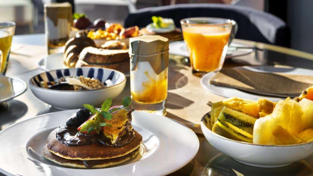 Uma mesa de café da manhã com panquecas, frutas, suco, granola, iogurte e torradas, servidos em pratos e copos brancos, servido no Vitoia Stone Hotel.