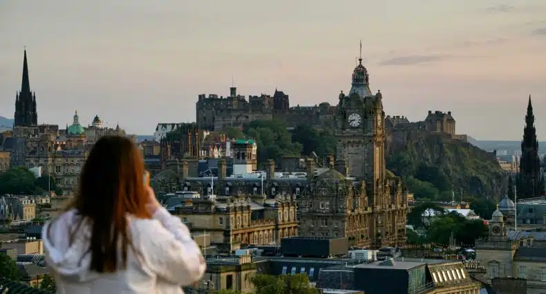Uma pessoa observa a paisagem urbana de Edimburgo, com edifícios históricos e o Castelo de Edimburgo visíveis ao fundo ao anoitecer. Ilustra o eSIM Escócia.