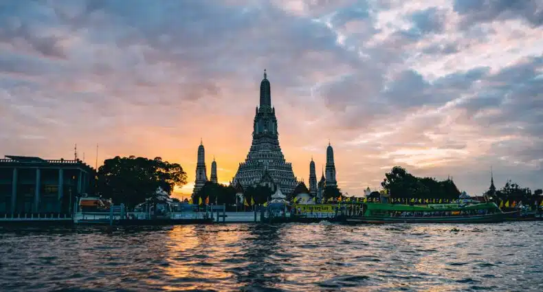 Um templo à beira do rio com torres altas é visto em silhueta ao pôr do sol, com barcos e água em primeiro plano, em Bangkok. Representa eSIM Bangkok.