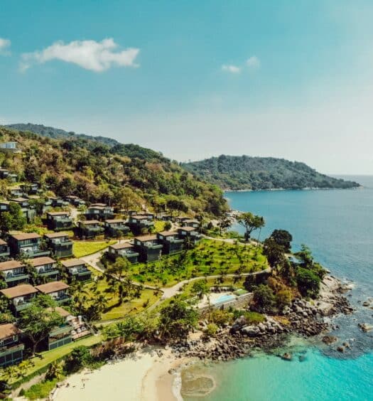 Vista aérea de vilas à beira-mar em uma encosta exuberante com vista para o oceano azul cristalino e uma praia de areia com litoral rochoso.