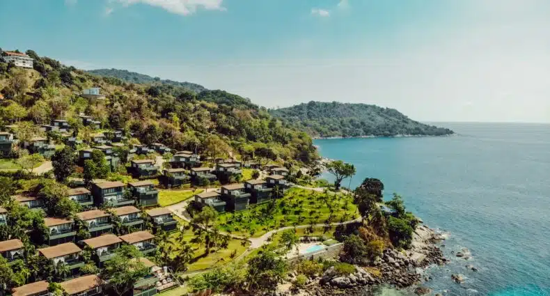 Vista aérea de vilas à beira-mar em uma encosta exuberante com vista para o oceano azul cristalino e uma praia de areia com litoral rochoso.