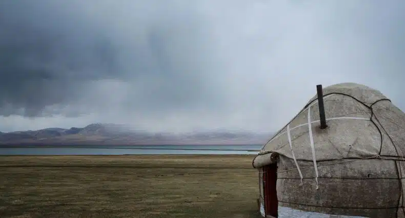 Uma yurt redonda fica em uma planície gramada perto de um lago, com montanhas e um céu nublado ao fundo, em Quirguistão . Representa eSIM Quirguistão.