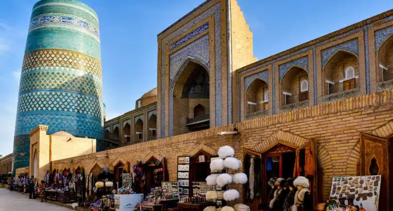 Vendedores vendem souvenirs e chapéus ao longo de uma rua de tijolos ao lado de um minarete alto com azulejos azuis e um edifício histórico, em Khiva. Representa eSIM Uzbequistão.