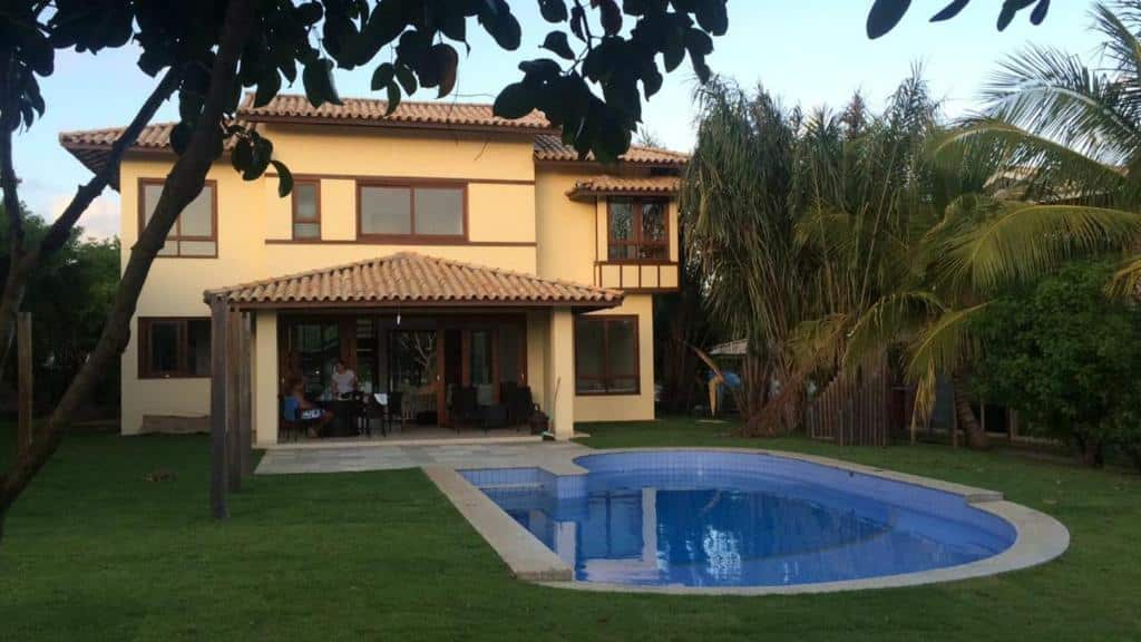 Casa bege de dois andares com telhado de telha, um pátio e uma piscina curva em um quintal gramado com palmeiras, trata-se da propriedade Casa de praia na Costa do Sauípe.