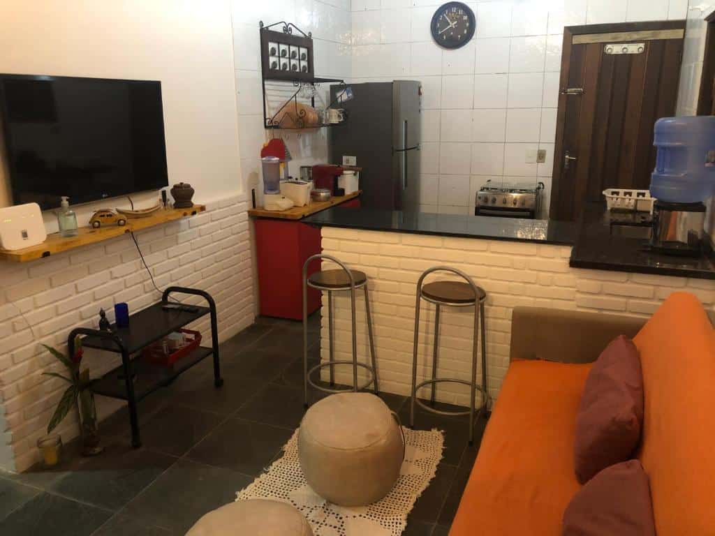 Uma pequena cozinha e sala de estar com sofá, banquetas, TV, geladeira, bebedouro e paredes de tijolos brancos, na Casa de praia em Paúba perto de cachoeira.