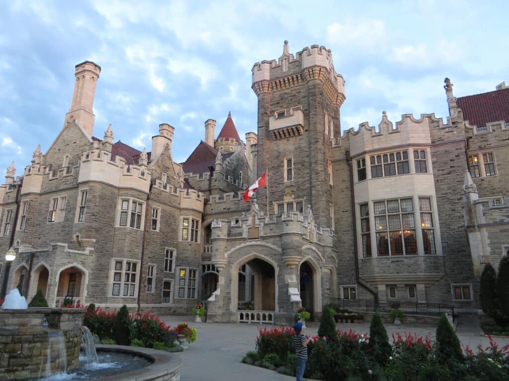 Um grande castelo de pedra com entrada em arco, torres e uma bandeira canadense, cercado por jardins e uma fonte.